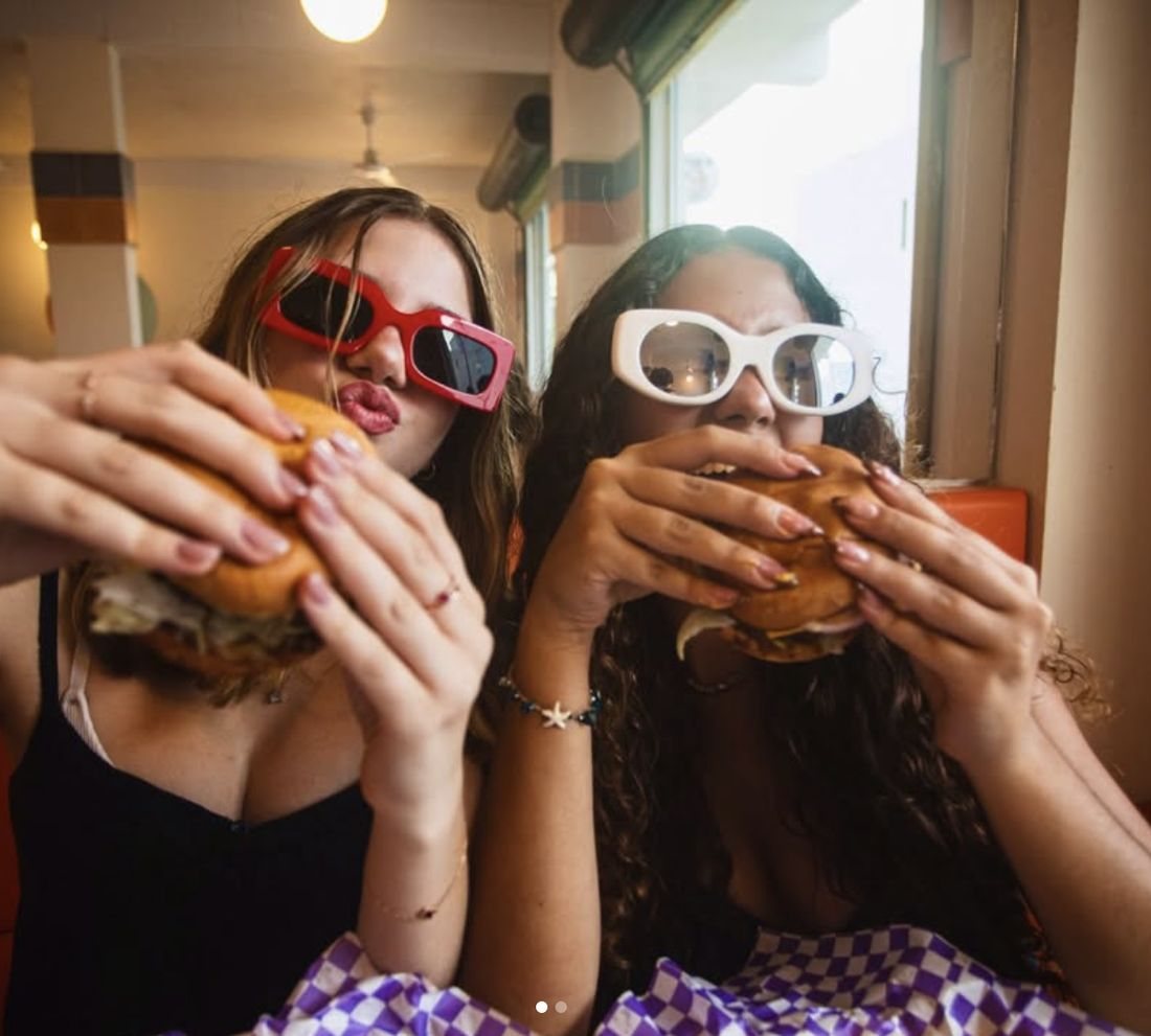 Dos amigas con lentes de sol mordiendo sus smash burgers adentro de El Bajón.